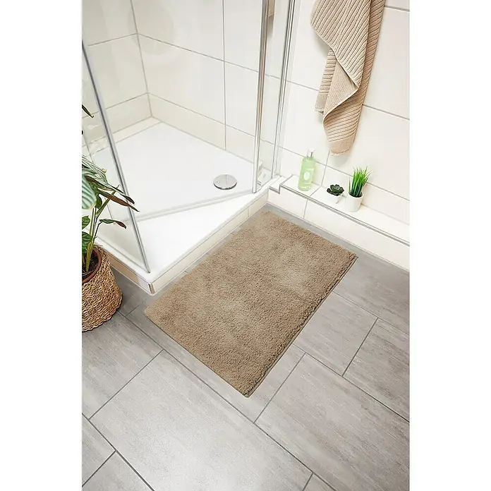 Badteppich Ibiza - 60 x 100 cm, Taupe, 100% Baumwolle - Ein flauschiger, beigefarbener Badteppich liegt vor einer Dusche mit weißen Fliesen und einer Zimmerpflanze daneben.