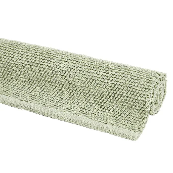 Badteppich Atlanta - 60 x 100 cm, Salbei, 100% Baumwolle - Ein gerollter, hellgrüner Badteppich mit Noppenstruktur liegt auf einem schwarzen Untergrund. Weiche Oberfläche für Komfort und Stil im Badezimmer.