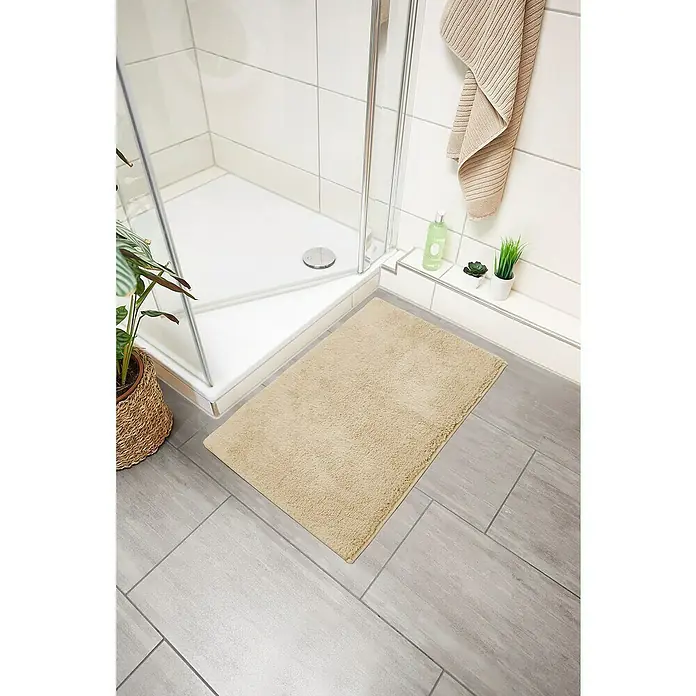 Badteppich Ibiza - 50 x 80 cm, Beige, 100% Baumwolle - Ein rechteckiger, beiger Badvorleger liegt vor einer Dusche mit Pflanzen und Handtuch, der auf grauen Fliesen für einen warmen Akzent sorgt.