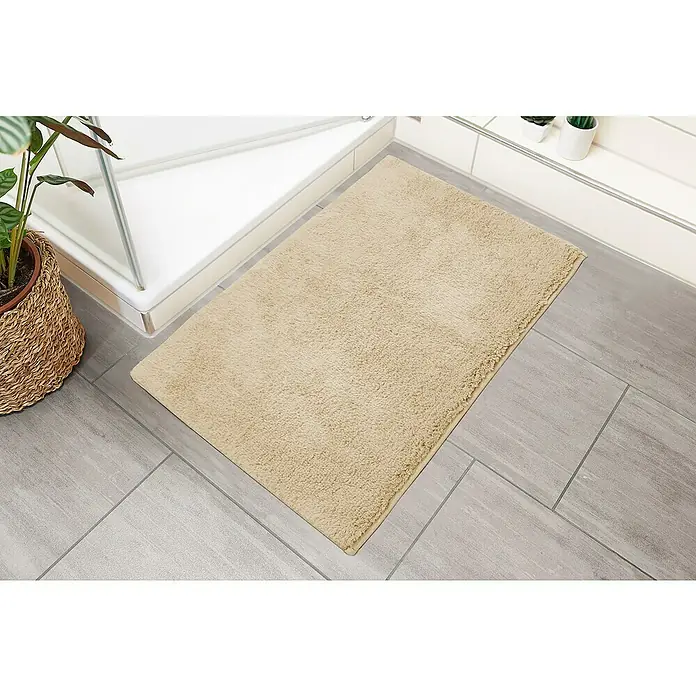 Badteppich Ibiza - 50 x 80 cm, Beige, 100% Baumwolle - Beige Badematte liegt auf grauen Fliesen neben einer Duschkabine und einem Korb mit einer Zimmerpflanze, sorgt für Wärme und Komfort.