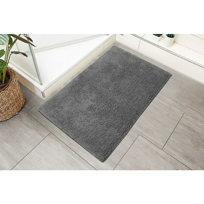 Badteppich Ibiza - 60 x 100 cm, Anthrazit, 100% Baumwolle - Weicher, rechteckiger, grauer Badteppich auf hellgrauen Fliesen vor einer Duschkabine und Topfpflanzen.