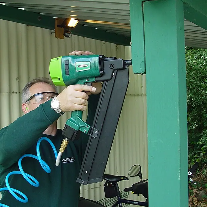 Prebena Druckluftnagler 7XR-RK90 - Betriebsdruck: 4 bar - 7 bar, Passend für: Prebena Rundkopf-Streifennägel der Type von RK 50 - 90 mm - Ein Mann benutzt einen PREBENA Druckluftnagler, um Holz zu befestigen. Der Nagler ist grün und schwarz und wird mit Druckluft betrieben.