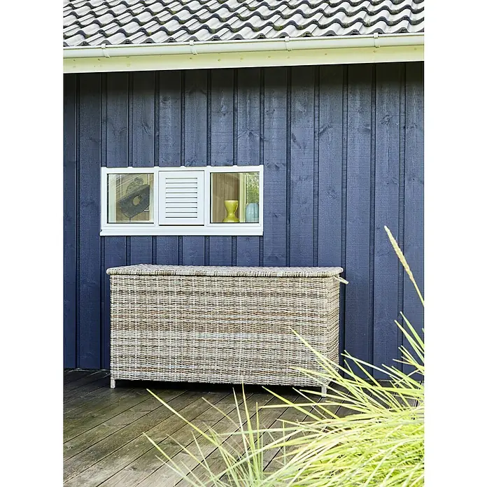 Baúl para jardín Gotland - 150 x 65 x 68 cm, Plástico, Arena - Vista exterior de una caja de almacenamiento rectangular de cesto de ratán sobre una terraza de madera frente a una pared de madera azul oscuro con ventana.