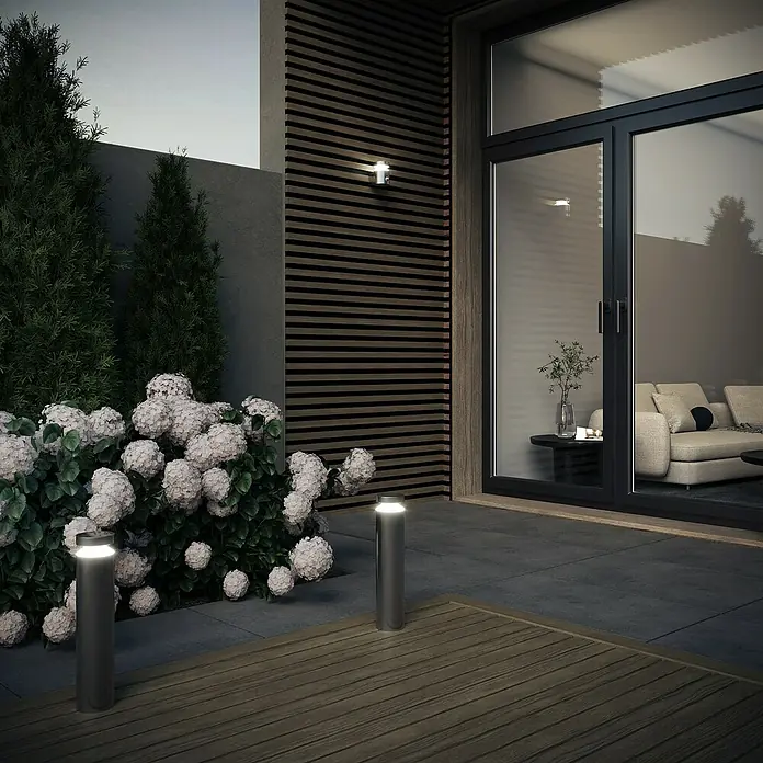 Osram Solar-Außenwandleuchte Wall - 6 W, L x B x H: 15 x 10,2 x 13,5 cm, IP44, 360 lm - Moderne Gartenbeleuchtung: Zwei zylindrische Außenleuchten beleuchten Blumen und eine Terrasse vor einem Haus mit Schiebetür.