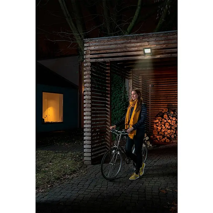 Osram LED-Strahler Endura Flood - 50 W, Anthrazit, Warmweiß, IP65 - Frau mit Fahrrad unter Außenleuchte. Diese LED-Leuchte sorgt für Sicherheit und Komfort im Außenbereich bei Nacht.