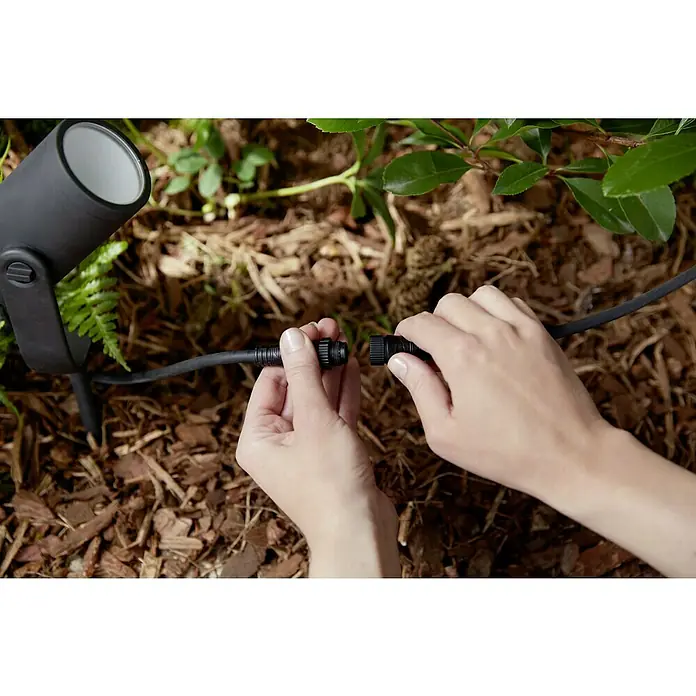 Philips Hue LED-Gartenspot Lily - 8 W, Lichtfarbe: RGB, IP65 - Nahaufnahme einer Person, die im Garten ein Erdkabel an eine schwarze Gartenleuchte anschließt.