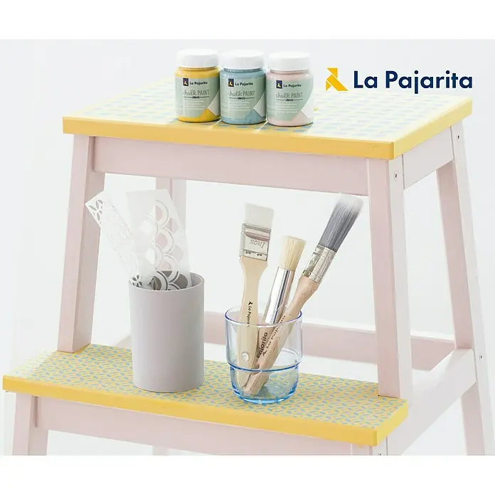 La Pajarita Pintura de tiza Chalk Paint - Vintage, 175 ml, Mate - Una foto de pinturas de tiza La Pajarita en amarillo, azul y rosa expuestas en un taburete decorado, junto con pinceles y plantillas.