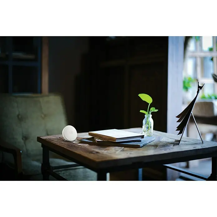 Airthings Luftqualitätsmonitor Wave Mini - Durchmesser: 80 mm, Batterielaufzeit: 3 Jahre - Weißes, rundes Smart-Home-Gerät auf einem Holztisch in einem gemütlichen Wohnzimmer. Das Gerät ist kompakt und unauffällig.