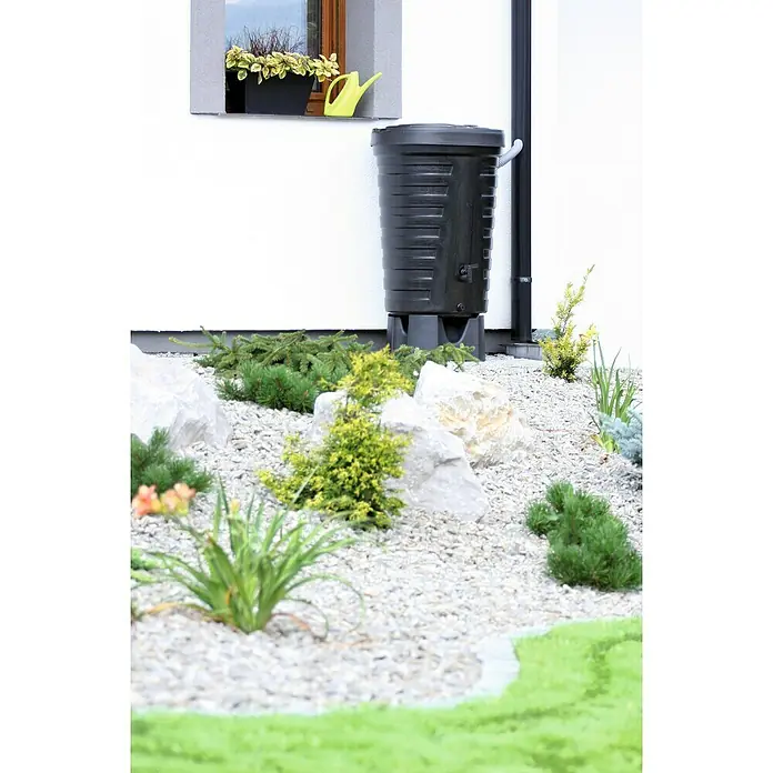 Regentonne Raincan - 210 l, Schwarz, 1 Stk. - Ein schwarzer Regenwassertank vor einer weißen Hauswand mit Kies und Pflanzen im Vordergrund.