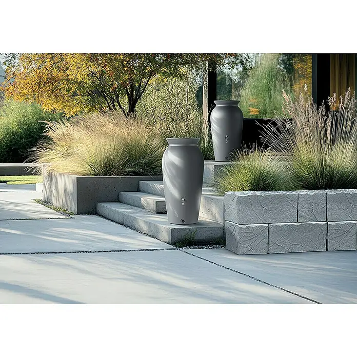 Regentonne Velcan - 120 l, Anthrazit - Zwei graue Wasserspeichergefäße im Garten, eingebettet in üppige Gräser und moderne Betonarchitektur. Funktionale Gartendekoration.