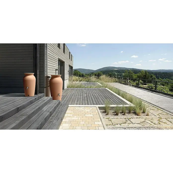 Regentonne Velcan - 230 l, Terrakotta - Zwei Terrakotta-Regentonnen mit Hähnen stehen auf einer Holzterrasse vor einem modernen Haus mit Blick auf eine grüne Hügellandschaft.