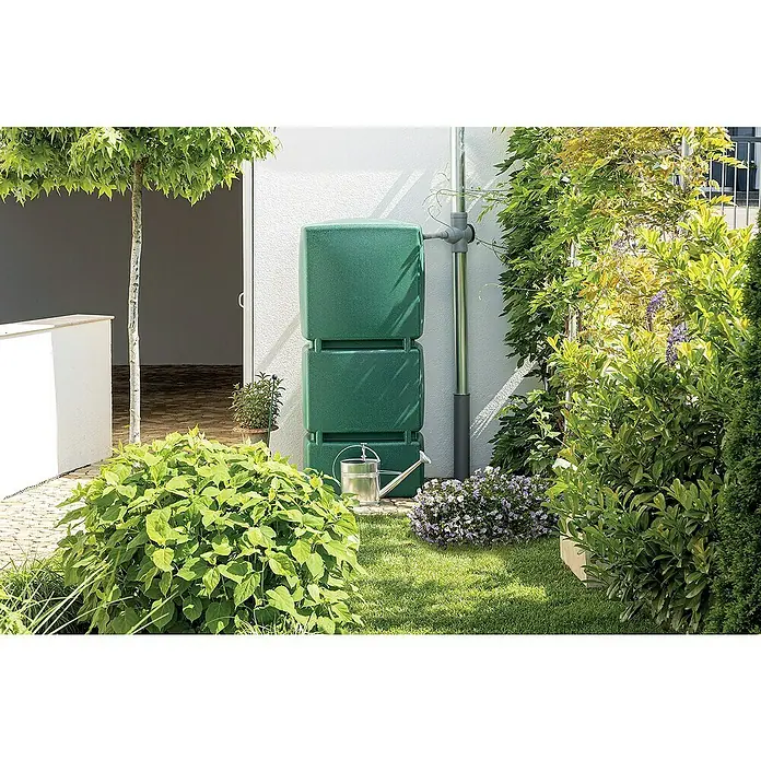 3P Technik Regentonne Jumbo - 800 l, Waldgrün - Gartenszene mit einem grünen Regenwassertank, der an einer weißen Wand steht, umgeben von Pflanzen und einem silbernen Gießkanne.