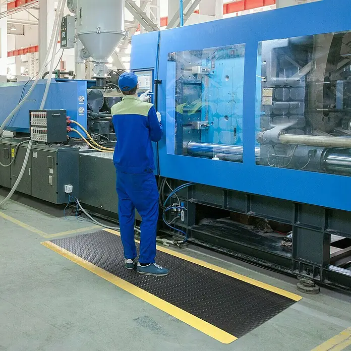Floordirekt Bodenschutzmatte Heavy-Duty Diamond - 90 x 150 cm, Gelb/Schwarz - Ein Arbeiter steht auf einer schwarzen Antirutschmatte mit gelben Rändern vor einer großen blauen Industriemaschine.