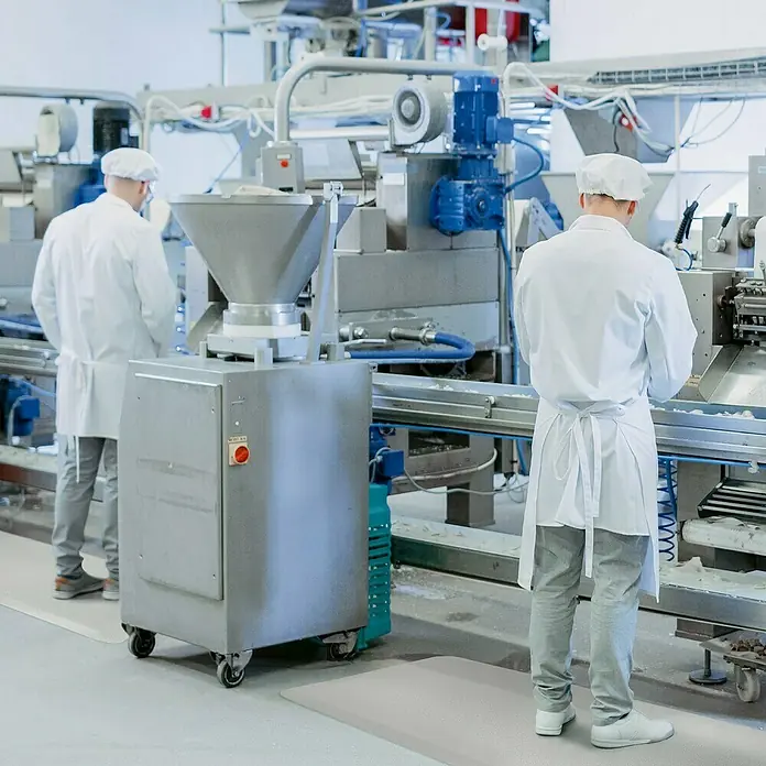 Floordirekt Bodenschutzmatte Soft-Tritt - 120 x 1.700 cm, Grau - Foto von zwei Arbeitern in steriler Kleidung neben einer Maschine, die als Lebensmittelverarbeitungsanlage verwendet wird.