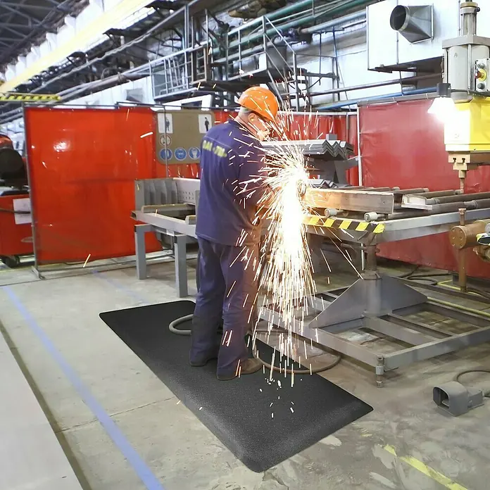 Floordirekt Bodenschutzmatte Heavy-Duty Soft - 60 x 90 cm, Schwarz - Arbeitsplatzmatte im Einsatz: Ein Arbeiter steht auf einer schwarzen, strapazierfähigen Antirutschmatte bei der Arbeit in einer Werkshalle.