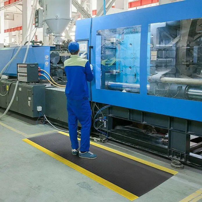 Floordirekt Läufer Softer-Work-Mat - 120 x 50 cm, Gelb/Schwarz - Ein Arbeiter steht auf einer schwarzen Anti-Ermüdungsmatte mit gelben Rändern vor einer großen blauen Industriemaschine in einer Fabrik.