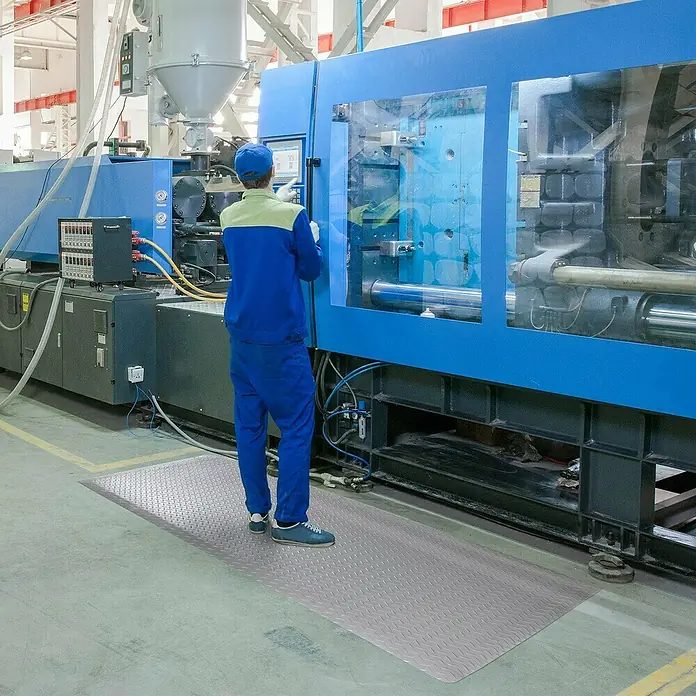 Floordirekt Bodenschutzmatte Heavy-Duty Diamond - 60 x 90 cm, Grau - Ein Arbeiter steht auf einer grauen Anti-Ermüdungsmatte vor einer großen blauen Maschine in einer Fabrik.