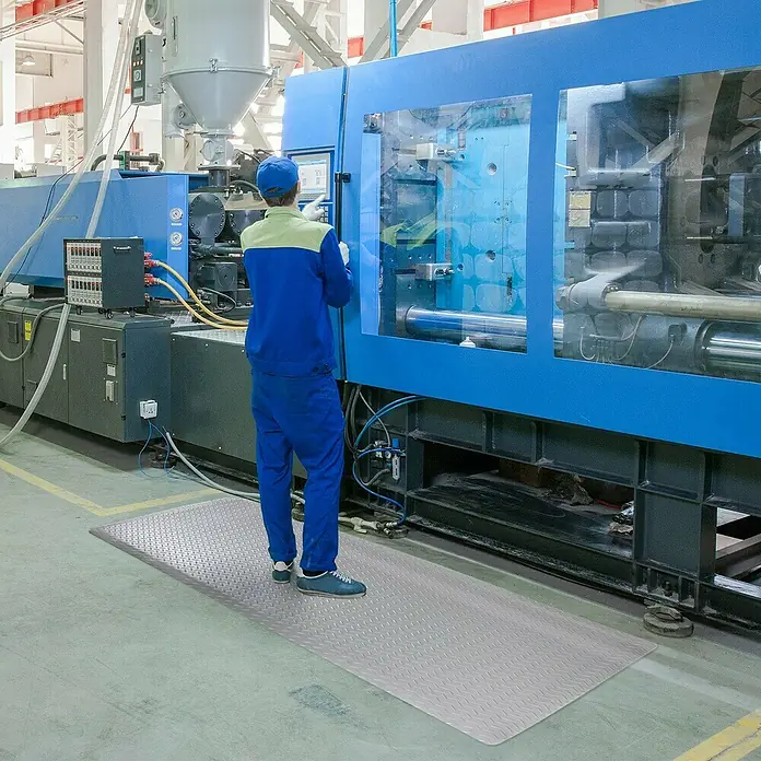 Floordirekt Fußbodenschutzmatte Dyna-Protect Diamond - Grau, 120 x 900 cm - Graue, geriffelte Antirutschmatte vor einer großen blauen Industriemaschine, die für Sicherheit am Arbeitsplatz sorgt.