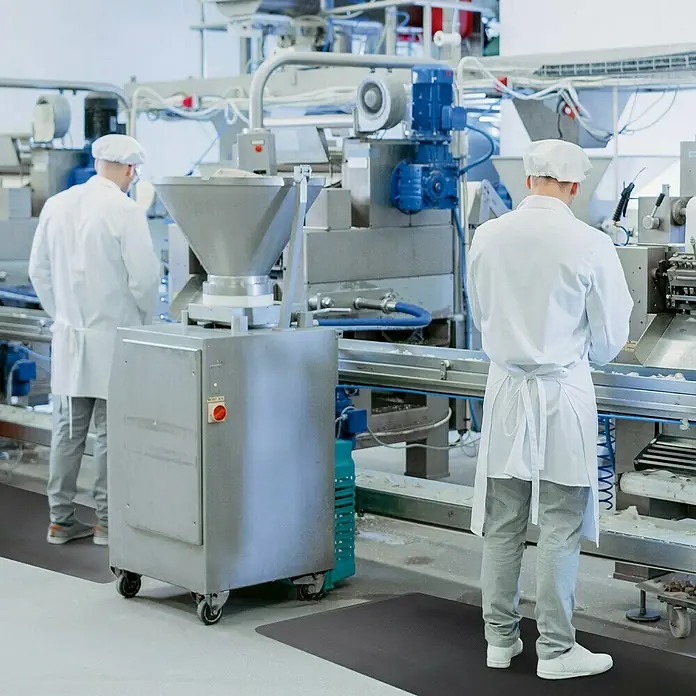 Floordirekt Bodenschutzmatte Heavy-Duty Ripp - 60 x 90 cm, Schwarz - Bild von zwei Personen, die in einer Fabrik auf einer schwarzen, ergonomischen Antirutschmatte stehen.\n