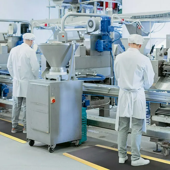 Floordirekt Bodenschutzmatte Heavy-Duty Ripp - 90 x 150 cm, Gelb/Schwarz - Bild von zwei Personen in Schutzkleidung, die in einer Fabrik auf schwarzen Matten mit gelben Rändern arbeiten.
