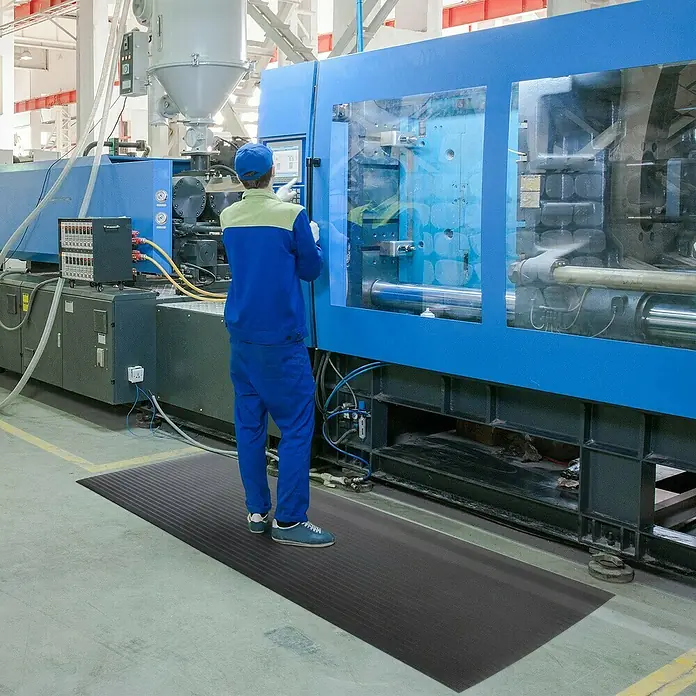 Floordirekt Läufer Softer-Work-Mat - 120 x 800 cm, Schwarz - Ein schwarze Anti-Ermüdungsmatte auf dem Boden vor einer blauen Maschine, mit einem Arbeiter, der davor steht.