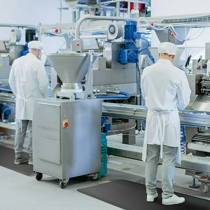 Floordirekt Bodenschutzmatte Soft-Tritt - 60 x 1.830 cm, Schwarz - Eine Fabrikhalle mit zwei Mitarbeitern, die auf einer schwarzen Antiverschleißmatte stehen, die vor Arbeitsplatzermüdung schützt.