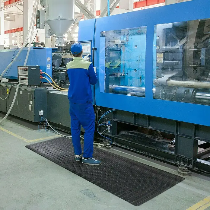 Floordirekt Fußbodenschutzmatte Dyna-Protect Diamond - Schwarz, 90 x 50 cm - Ein Arbeiter steht auf einer schwarzen Anti-Ermüdungsmatte vor einer großen Industriemaschine in einer Fabrik.