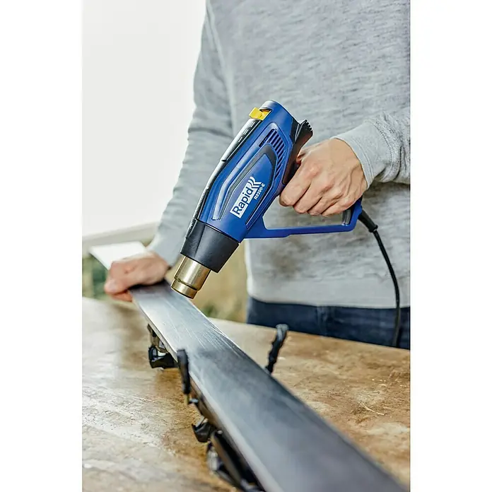 Rapid Heißluftgebläse R2200-E - 2.200 W, Temperaturbereich: 60 °C - 650 °C - Das Rapid R2200-E Heißluftgebläse wird zur Bearbeitung eines Skis verwendet. Das Gerät ist blau und wird mit einer Hand gehalten.