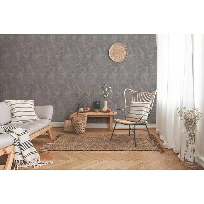 A.S. Création New Walls Vliestapete Dschungel - Taupe, Floral, 10,05 x 0,53 m - Innenaufnahme eines Raumes mit einem Rattansessel und einem kleinen Holztisch, der eine gemütliche Atmosphäre erzeugt.