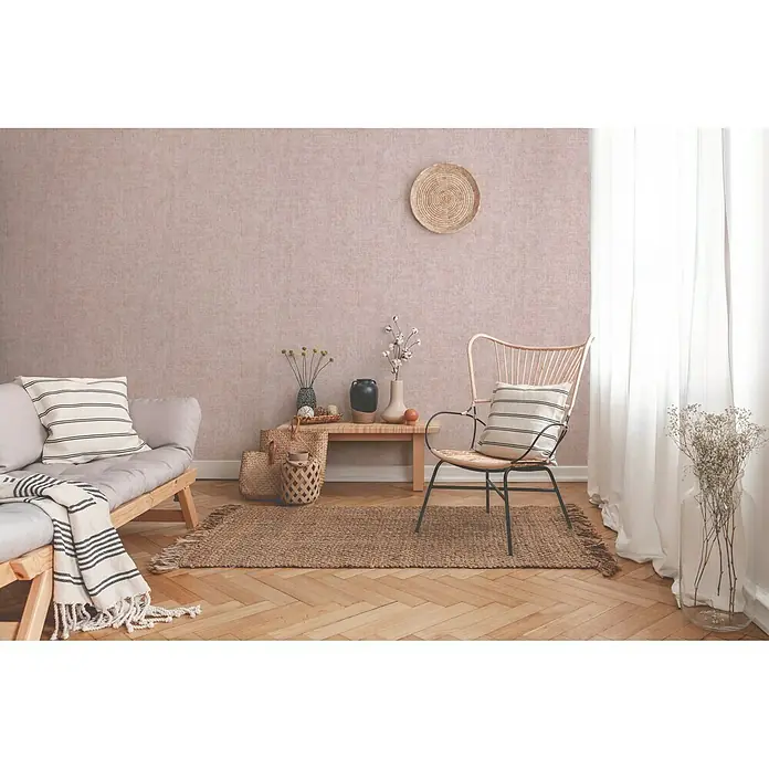 A.S. Création New Walls Vliestapete I - Rosa, Uni, 10,05 x 0,53 m - Das Bild zeigt ein stilvolles Wohnzimmer mit einem Korbsessel, einem Sofa mit Kissen und einem Teppich auf einem Holzboden.