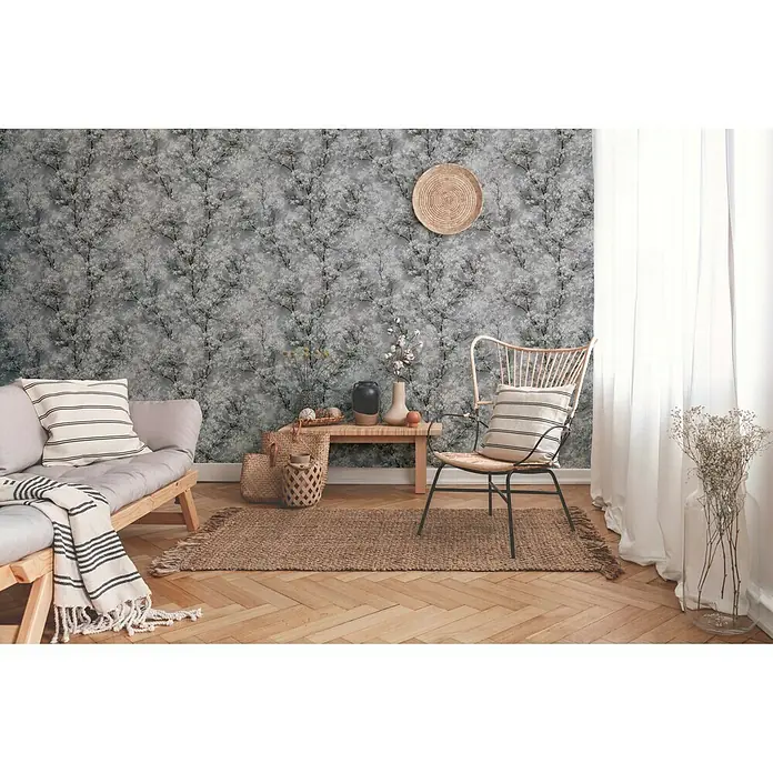 A.S. Création New Walls Vliestapete Bäume - Grau, Floral, 10,05 x 0,53 m - Zimmer mit Rattan-Stuhl und Couch. Der Rattan-Stuhl hat ein helles Holzgestell und ein dunkles Metalluntergestell mit einer gestreiften Kissenauflage.