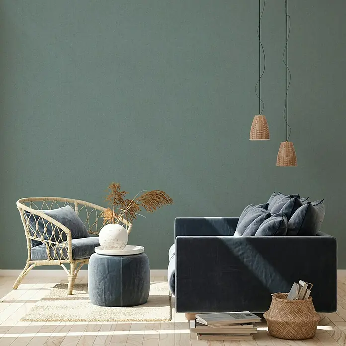 A.S. Création New Walls Vliestapete - Dunkelgrün, Uni, 10,05 x 0,53 m - Einladendes Wohnzimmer-Arrangement mit einem blauen Sessel, einem Korbsessel und dekorativen Elementen. Die Hängelampen verleihen dem Raum eine warme Note.