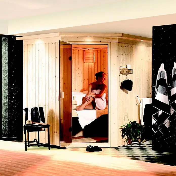 Karibu Elementsauna Lilja - Ohne Ofen, Ohne Dachkranz, L x B x H: 196 x 170 x 192 cm - Eine Frau entspannt in einer hell erleuchteten Holzsauna. Handtücher und eine Pflanze schmücken den Raum.