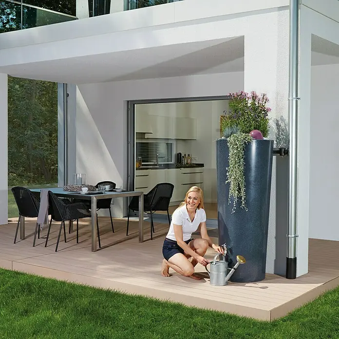 Garantia Modern Line Regenspeicher Stone 2 in 1 - 350 l, Lava - Eine Frau befüllt eine Gießkanne an einem grauen Wasserspeicher mit Pflanzen, der an einem modernen Haus steht.