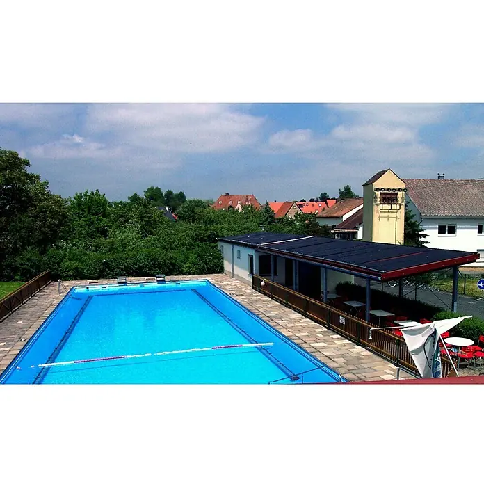 Sunset Solar-Schwimmbadanlage Sunstar 1 - Einsatzbereich: Schwimmbaderwärmung - Ein Freibad mit blauem Wasser, umgeben von Bäumen und Gebäuden, lädt zum Schwimmen und Entspannen im Sommer ein.