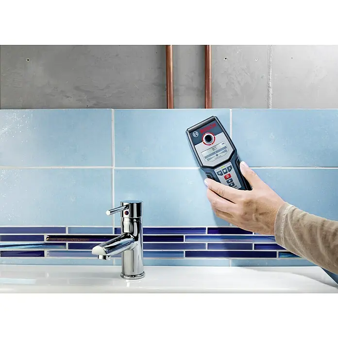 Bosch Professional Detector GMS 120 - Profundidad de detección: Máx. 120 mm de acero, Tiempo de funcionamiento: 5 h - Un escáner de pared Bosch sostenido contra azulejos azules, utilizado para detectar objetos ocultos. Perfecto para instalaciones seguras en baños y cocinas.