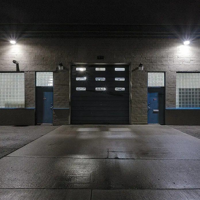 Arte confort Lusna Farola LED con soporte - 8 W, 700 lm, 6.500 K, IP54 - Una puerta de garaje industrial entre dos puertas azules con ladrillos de vidrio y brillantes focos superiores ilumina la noche.