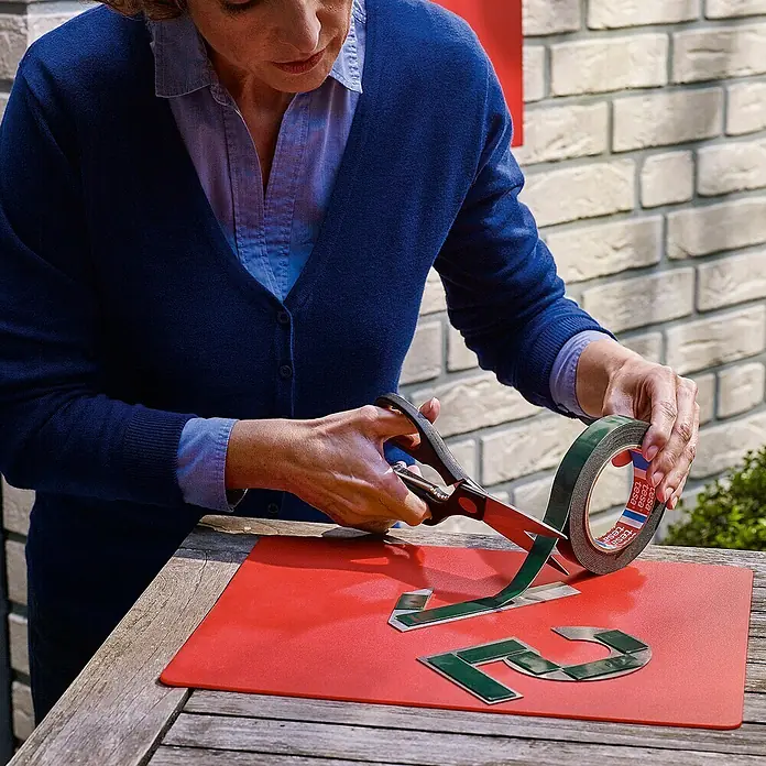 Tesa Montageband - Geeignet für: Mauerwerk, Belastbarkeit pro m: 10 kg, 1,5 m x 19 mm - Nahaufnahme einer Frau, die Tesa Montageband zuschneidet, um es auf einer roten Matte zu befestigen. Das graue Band ist eine großartige Lösung.