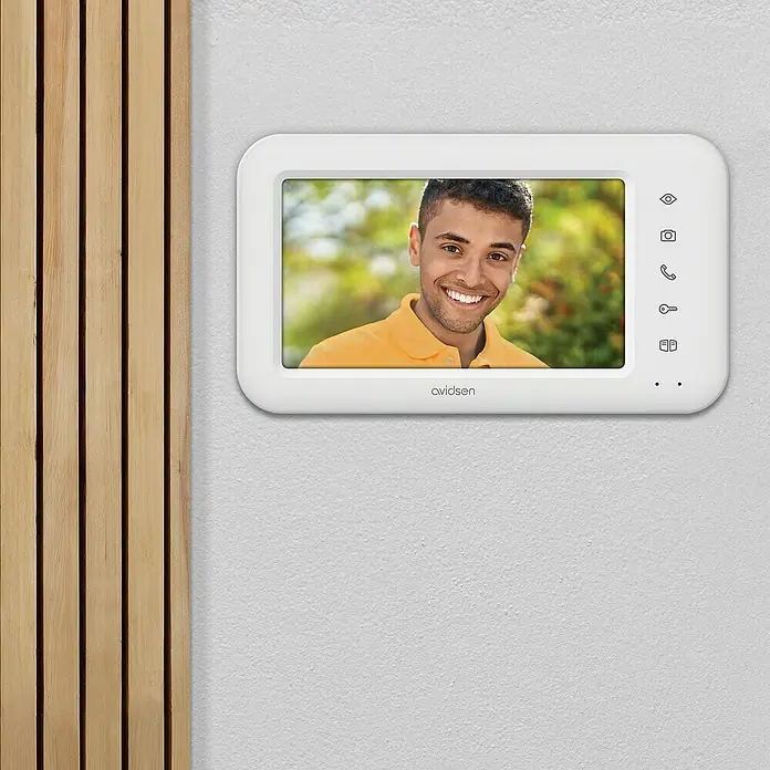 Avidsen Funk-Video-Türsprechanlage drahtlos mit Solarpanel und Batterie Elia Solar Wave - 125 ° - Avidsen Video-Türsprechanlage mit einem lächelnden Mann auf dem Display, an einer Wand montiert neben einer Holzverkleidung.