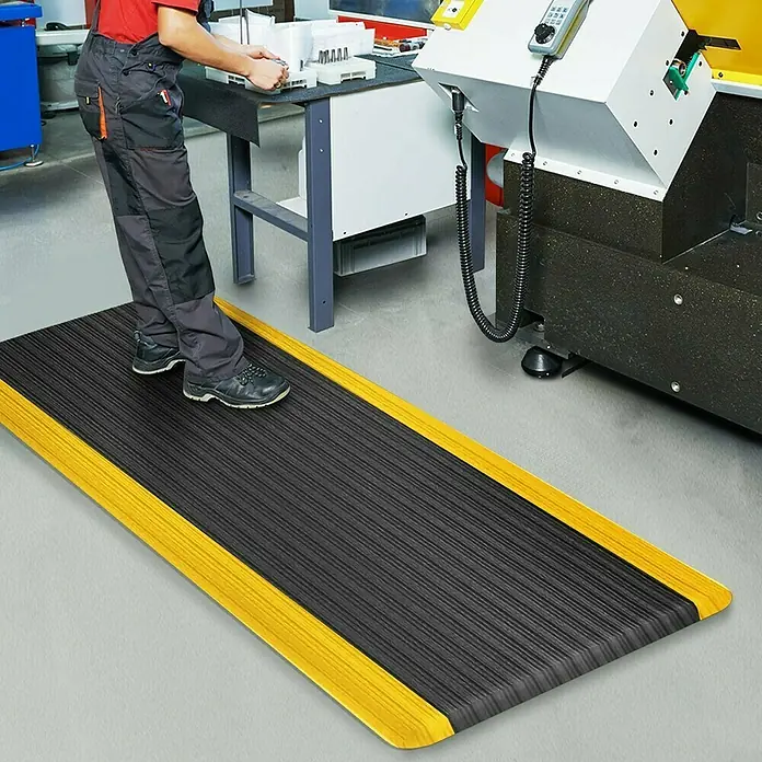 Floordirekt Bodenschutzmatte Heavy-Duty Ripp - 90 x 150 cm, Gelb/Schwarz - Eine schwarze, gerippte Antiverschmutzungsmatte mit gelben Rändern liegt auf dem Boden. Ein Mann steht auf der Matte in einer Fabrik.