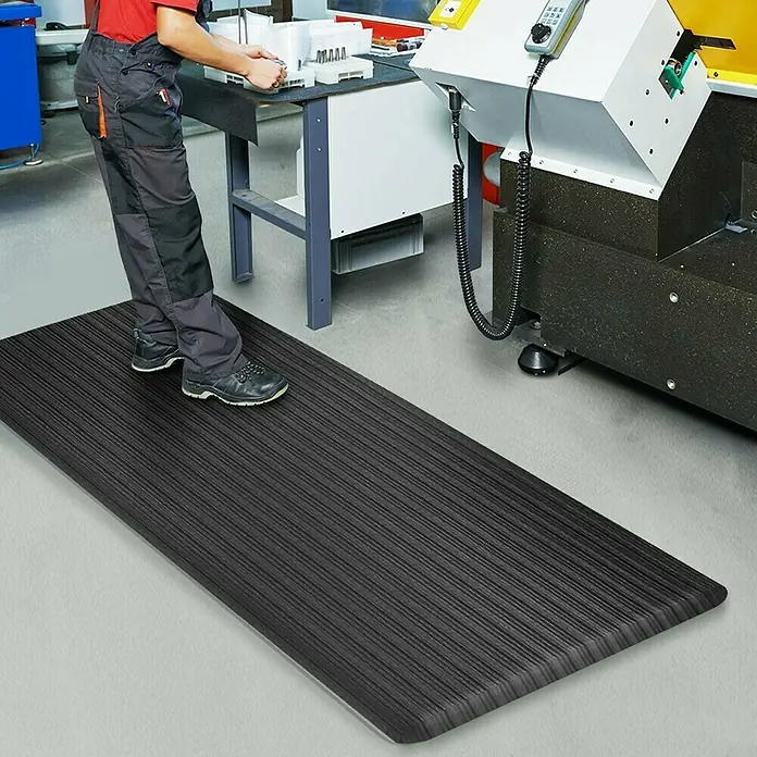 Floordirekt Bodenschutzmatte Heavy-Duty Ripp - 60 x 90 cm, Schwarz - Eine schwarze Anti-Ermüdungsmatte liegt auf dem Boden, eine Person steht darauf und arbeitet an einer Maschine.