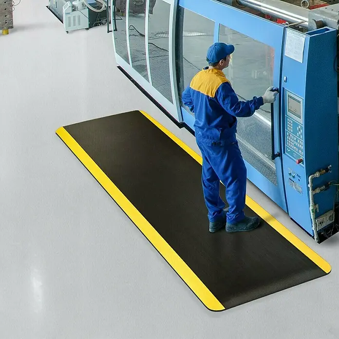 Floordirekt Bodenschutzmatte Soft-Tritt - 120 x 450 cm, Gelb/Schwarz - Ein Arbeiter steht auf einer schwarzen Anti-Ermüdungsmatte mit gelben Streifen vor einer Maschine in einer Fabrik.
