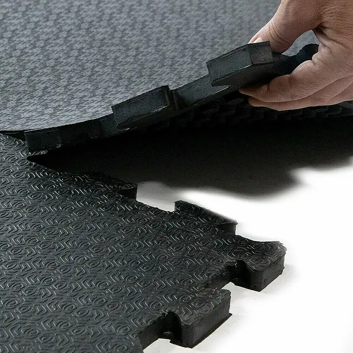 Floordirekt Läufer Puzzlev Lagama - 120 x 80 cm, Schwarz - Zwei schwarze, ineinandergreifende Schaumstoffmatten für Fitness und Sport, die von einer Hand gehalten werden.