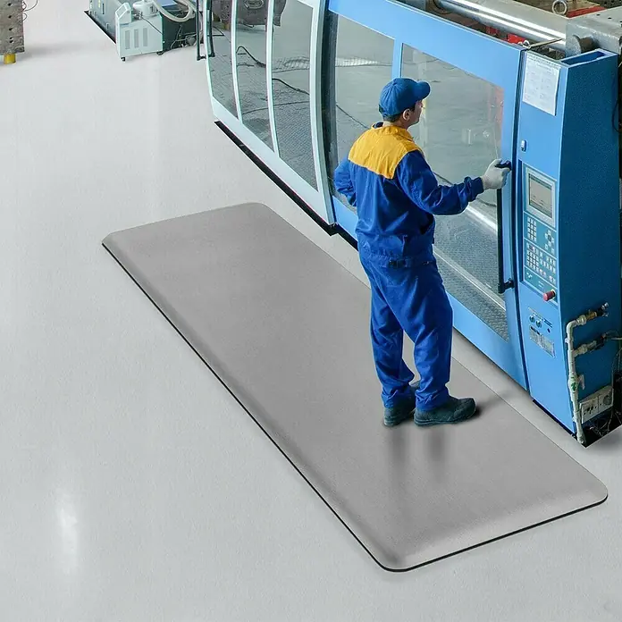 Floordirekt Bodenschutzmatte Soft-Tritt - 120 x 1.700 cm, Grau - Eine graue Antistressmatte liegt vor einer Werkzeugmaschine, auf der ein Arbeiter steht, um die Belastung durch langes Stehen zu reduzieren.