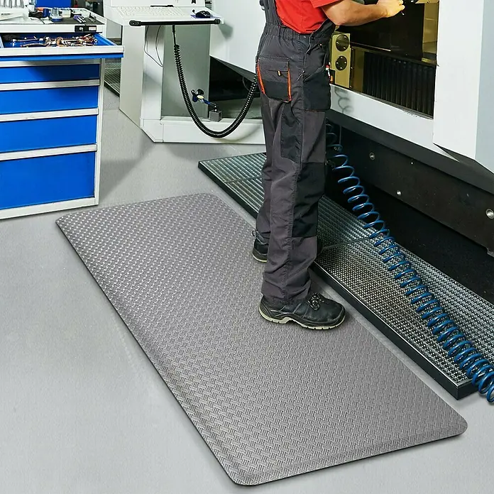 Floordirekt Bodenschutzmatte Heavy-Duty TireTuff - 90 x 150 cm, Grau - Ein Arbeiter steht auf einer grauen Anti-Ermüdungsmatte mit Rautenmuster vor einer Werkzeugmaschine.