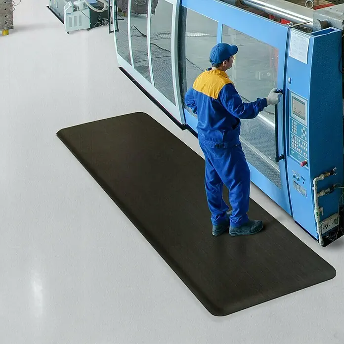 Floordirekt Bodenschutzmatte Soft-Tritt - 60 x 1.830 cm, Schwarz - Ein Mann steht auf einer schwarzen Anti-Ermüdungsmatte vor einer großen blauen Maschine. Ideal für Arbeitsplätze, die Komfort brauchen.