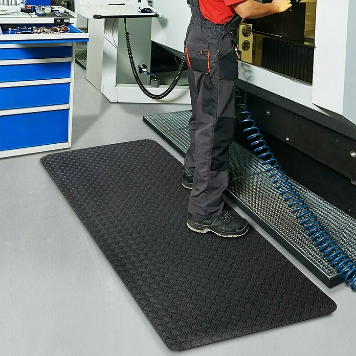 Floordirekt Bodenschutzmatte Heavy-Duty TireTuff - 90 x 150 cm, Schwarz - Hier ist eine rutschfeste, schwarze Bodenmatte mit Noppenmuster, die in einer Werkstatt vor einer Maschine liegt.