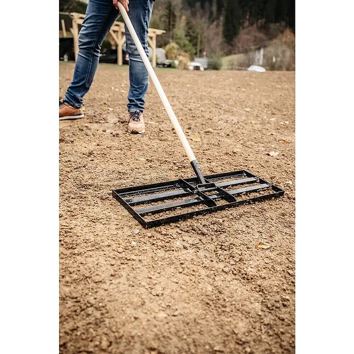 Gardol Rasenrakel ohne Stiel - 24,5 cm, Stahl, Passend für: Gerätestiele mit Konus, Ø 28 mm - Eine Person ebnet mit einem Bodengleichner die Erde, um sie für die Bepflanzung vorzubereiten. Das Werkzeug ist schwarz und hat einen Holzgriff.