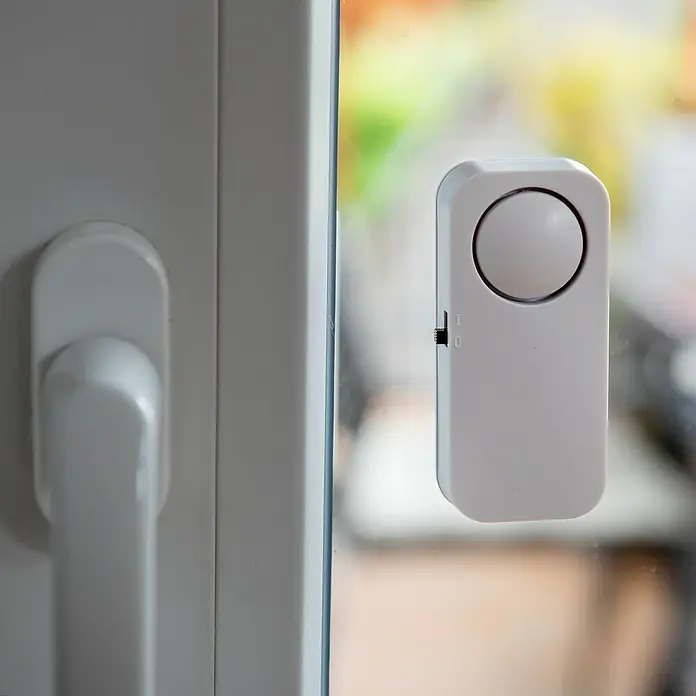 UniTEC Glasbruchmelder Glasbruchsensor - Weiß - Ein weißer Fenster- oder Türalarm ist an einem Fenster befestigt, mit einem Schalter an der Seite zur Steuerung der Aktivierung.
