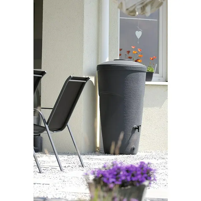 Regentonne Wallycan - 270 l, Anthrazit, 1 Stk. - Grauer Regenwassertank steht auf Kies, umgeben von Gartenstühlen und Blumen, und bietet eine nachhaltige Möglichkeit der Wassernutzung.